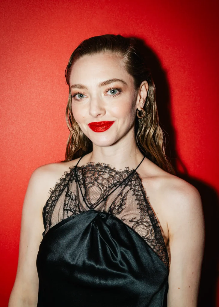 Portrait der Schauspielerin Amanda Seyfried vor einem roten Hintergrund, mit auffallend rotem Lippenstift und schwarzem, eleganten Spitzenkleid.