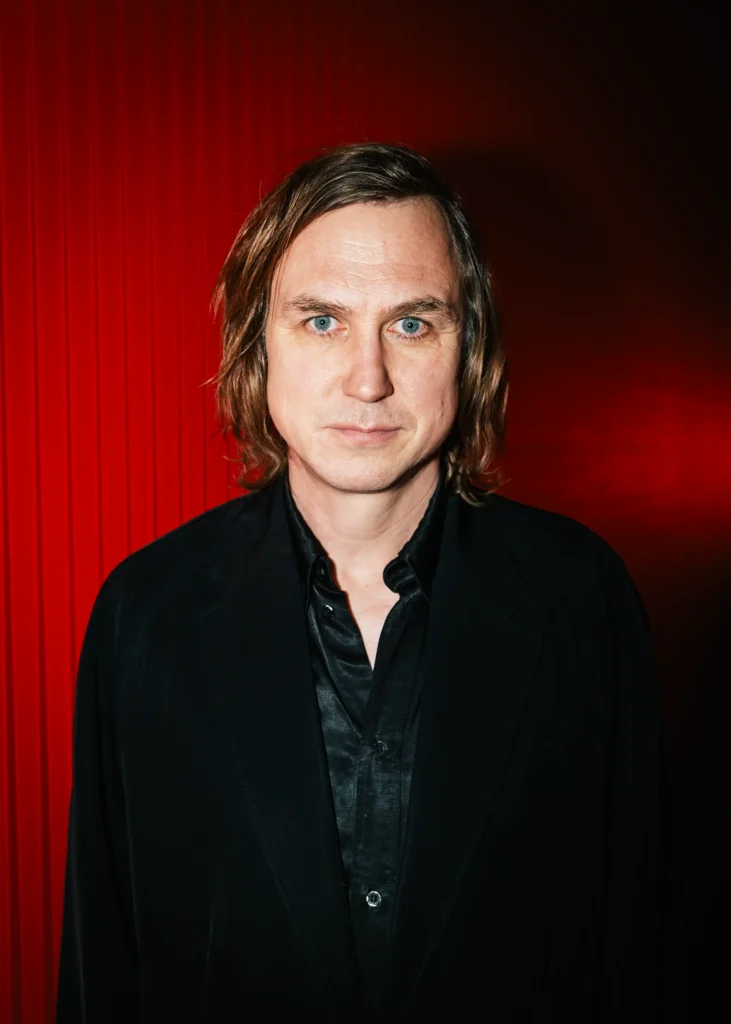 Portrait von Schauspieler Lars Eidinger vor intensiv rotem Hintergrund, frontal in die Kamera blickend.