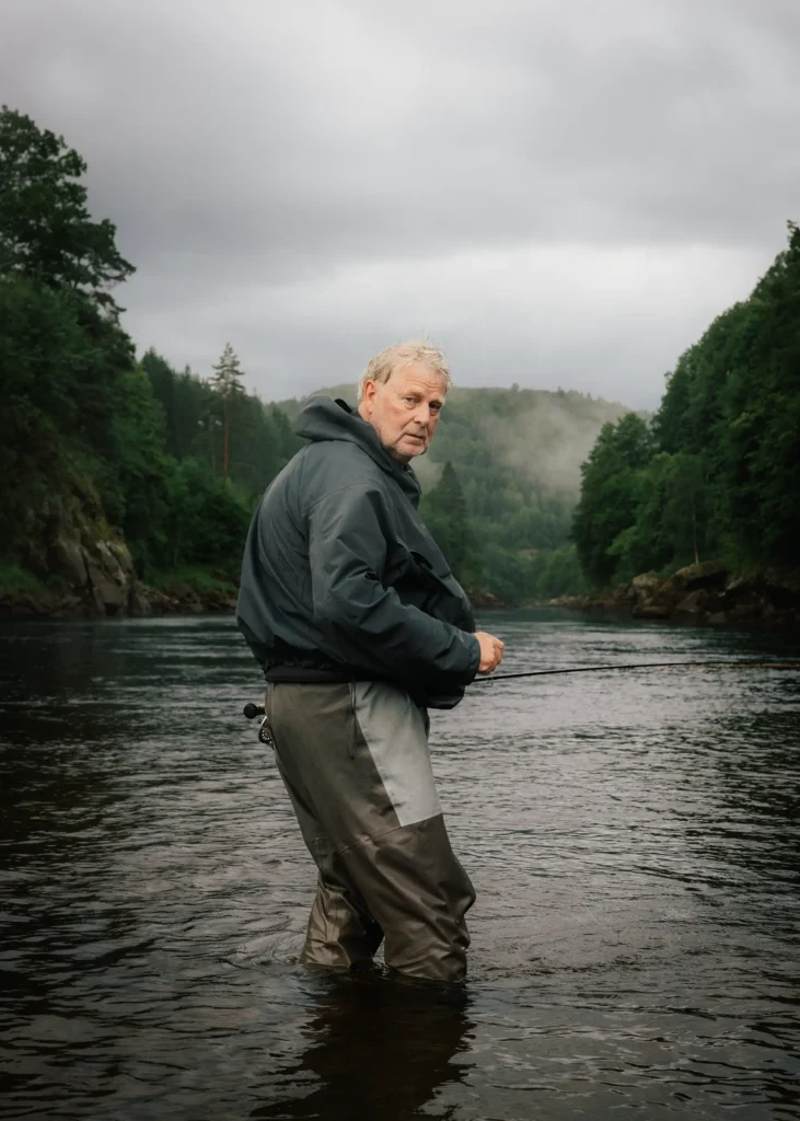 Das Bild zeigt einen älteren Mann in Anglerkleidung, der in einem Fluss steht und über die Schulter zur Kamera blickt. Er ist mit einer dunklen Jacke und Watthosen bekleidet, inmitten einer bewaldeten, nebligen Flusslandschaft unter einem bedeckten Himmel.